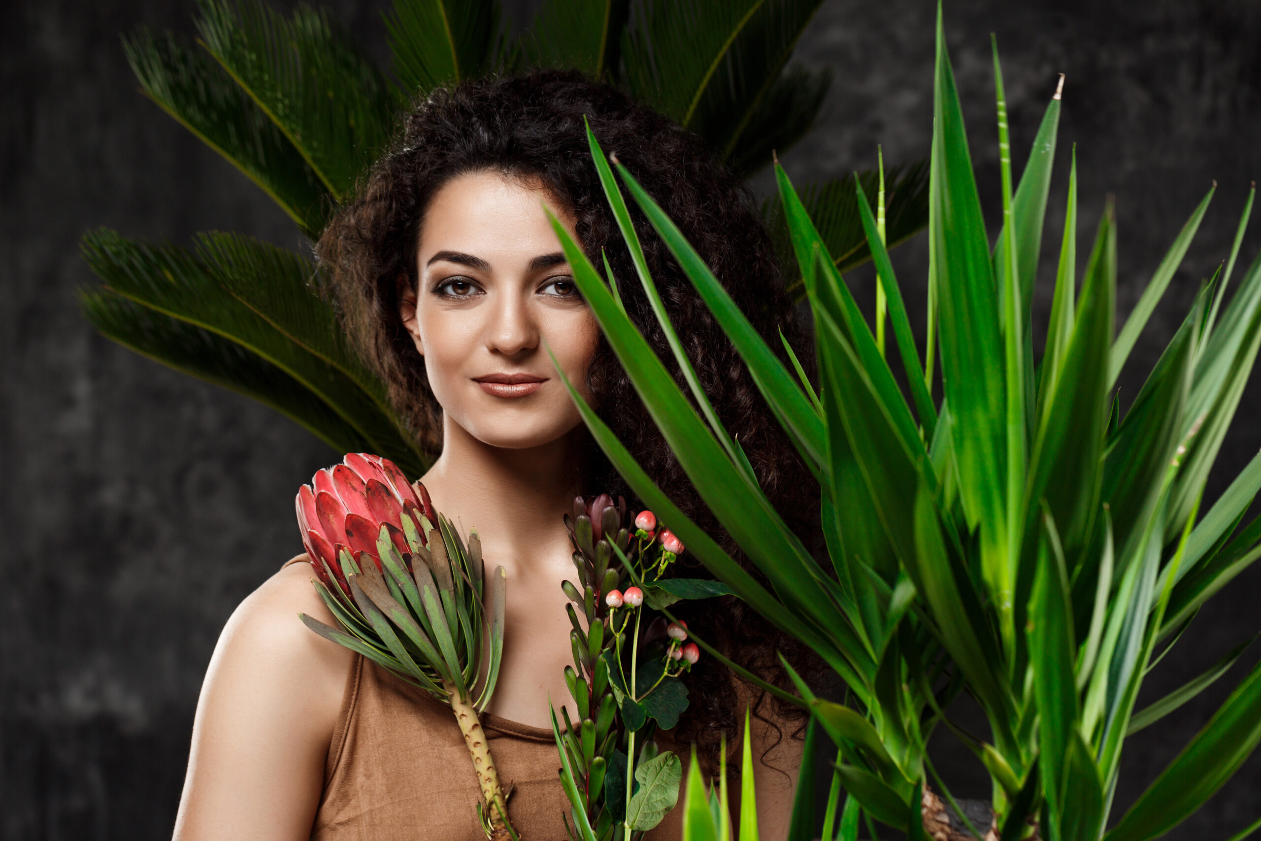 Young beautiful brunette girl in tropical plants looking at camera, smiling over grey background.